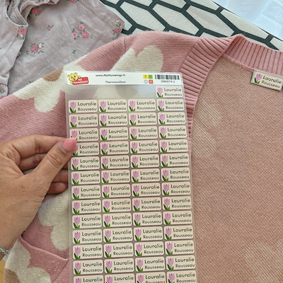 Main tenant une planche d'étiquettes thermocollantes au nom de Lauralie devant un gilet rose étiqueté